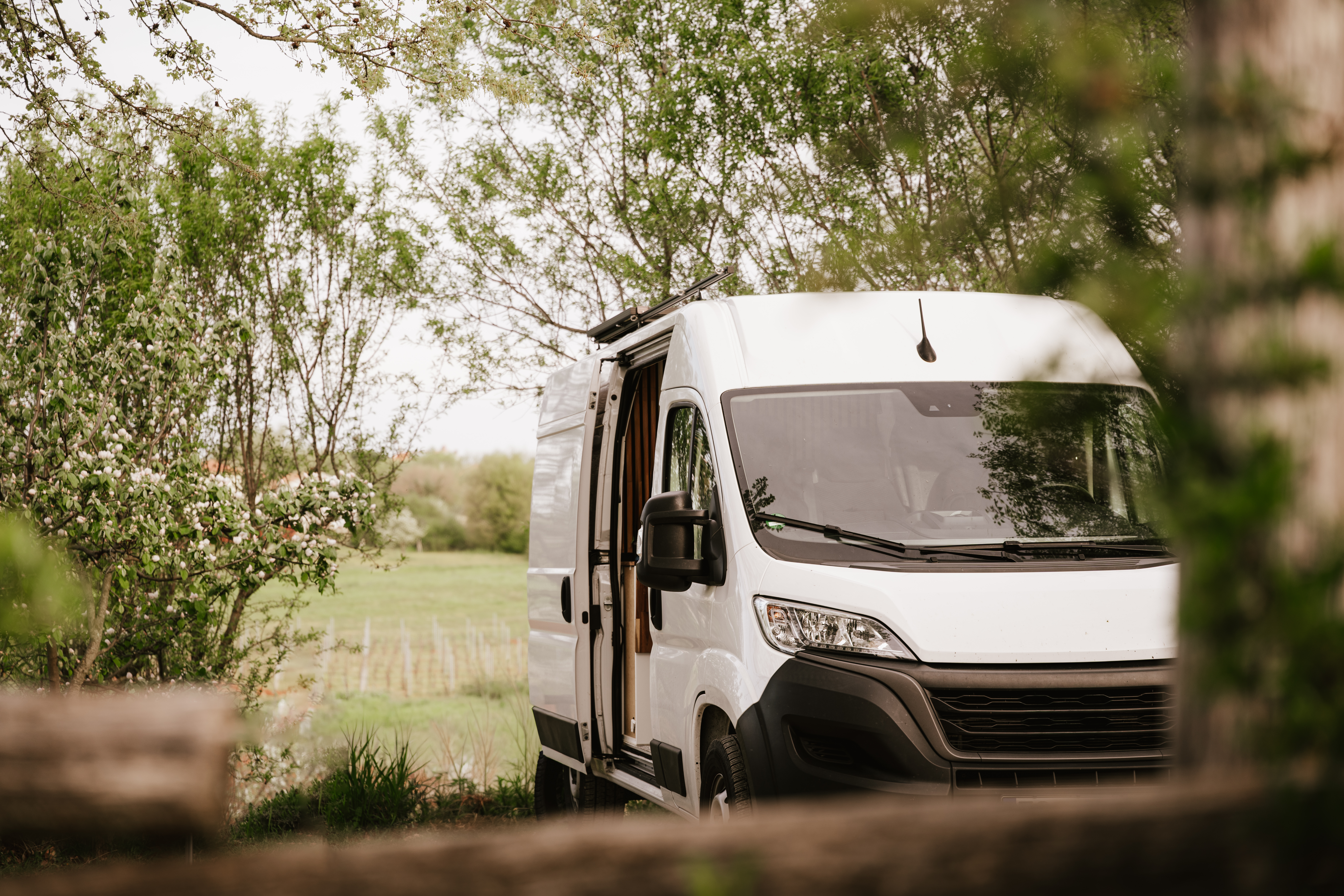 A modern white camper van parked peacefully in a green countryside setting, surrounded by blooming trees and soft natural light. Framed through rustic wooden elements and fresh spring foliage, the image creates a calm and inviting atmosphere of outdoor living and van life adventure. The open side door adds a sense of freedom, travel, and cozy mobile living. Perfect for themes related to camper lifestyle, road trips, remote work, nature retreats, sustainable travel, and minimalist outdoor living in spring.